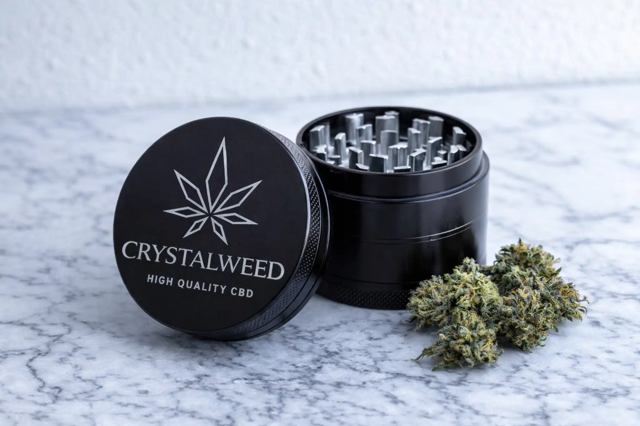 Grinder nero Crystalweed su piano in marmo con illuminazione fredda; coperchio con logo in primo piano, camera di macinatura aperta sullo sfondo e cime di cannabis accanto.